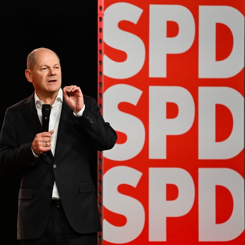 Scholz steht mit Mikro auf Bühne und spricht vor einer SPD-Werbewand.