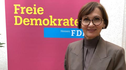 Eine Frau mit kurzen Haaren, Brille und grauem Blazer und grauer Bluse steht vor einer zimtroten Stellwand. Darauf steht in Gelb "Freie Demokraten FDP" 