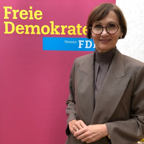 Eine Frau mit kurzen Haaren, Brille und grauem Blazer und grauer Bluse steht vor einer zimtroten Stellwand. Darauf steht in Gelb "Freie Demokraten FDP" 