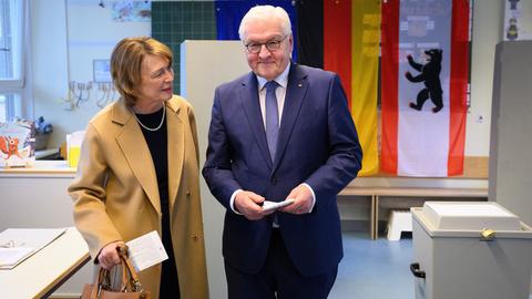 Ein Mann im blauen Anzug und eine Frau im beigen Mantel stehen nebeneinander in einem Raum. Beide halten Zettel in der Hand. Im Hintergrund sind die deutsche und Berliner Flagge zu sehen. Links im Bild stehen ein Tisch und ein Fenster, rechts eine Wahlurne.