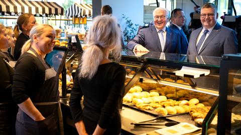 Bundespräsident Steinmeier, ein weißhaariger Mann, mit einem beleibten Mann an einem Bäckerstand auf einem Wochenmarkt. Die beiden Verkäuferinnen sind von hinten zu sehen. Die beiden Männer lachen.