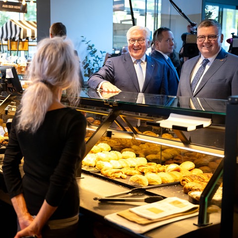 Bundespräsident Steinmeier, ein weißhaariger Mann, mit einem beleibten Mann an einem Bäckerstand auf einem Wochenmarkt. Die beiden Verkäuferinnen sind von hinten zu sehen. Die beiden Männer lachen.