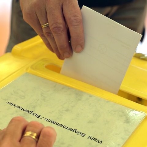 Wahlzettel wird in Wahlurne geworfen, mit Aufschrift: Wahl Bürgermeister/Bürgermeisterin