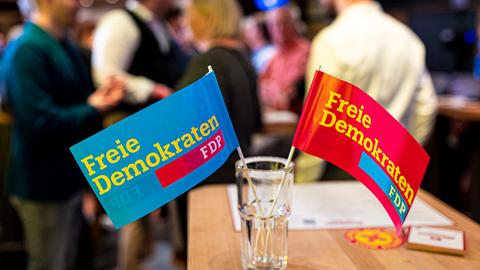 Nahaufnahme: Zwei Fähnchen mit der Aufschrift "Freie Demokraten FDP" stehen in einem Glas auf einem Tisch. Im Hintergrund unscharf stehende Menschen.