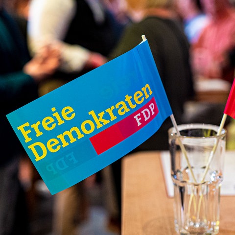 Nahaufnahme: Zwei Fähnchen mit der Aufschrift "Freie Demokraten FDP" stehen in einem Glas auf einem Tisch. Im Hintergrund unscharf stehende Menschen.