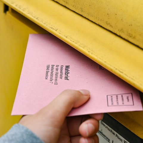 Eine Hand wirft einen rosa Briefwahlumschlag in den Schlitz eines gelben Briefkastens.