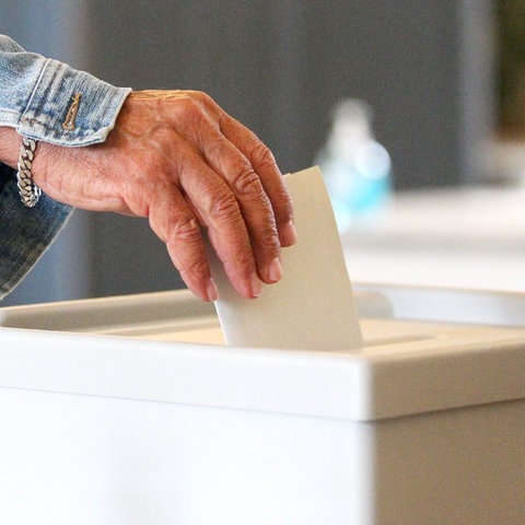 Eine Hand steckt einen Umschlag in eine versiegelte Wahlurne