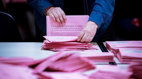 Zwei Hände halten rote Briefwahlbriefe (scharf im Bildhintergund). Im Bildvordergund unscharf viele rote Briefwahlbriefe ungeordnet auf einem Tisch.