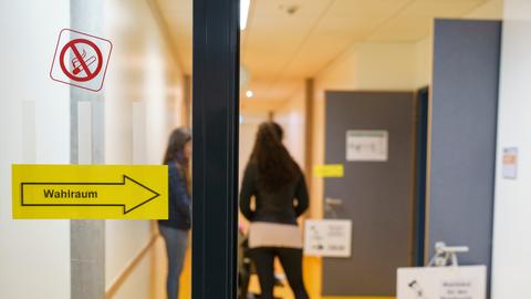 Glastür mit Schild "Wahlraum", dahinter Flur einer Schule, zwei verschwommene Personen sind zu erkennen