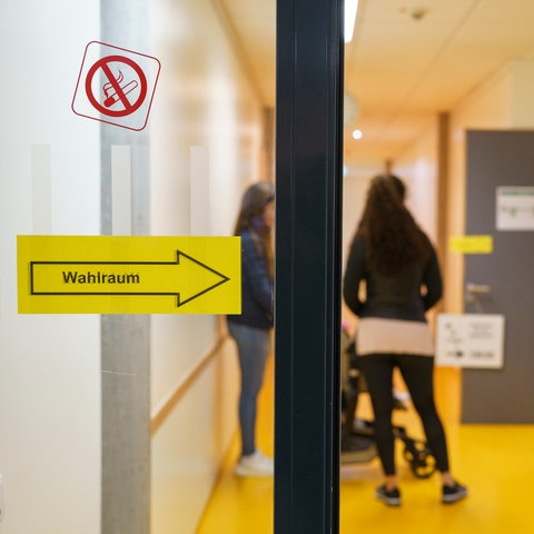 Glastür mit Schild "Wahlraum", dahinter Flur einer Schule, zwei verschwommene Personen sind zu erkennen