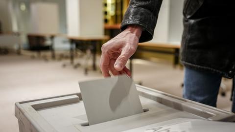 Eine Hand steckt einen Stimmzettel in eine Wahlurne. Die Person trägt eine schwarze Jacke. Im Hintergrund sind Tische und Wahlkabinen zu sehen. Ein Teil des Texts auf der Wahlurne ist sichtbar: „Abde...“.