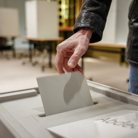 Eine Hand steckt einen Stimmzettel in eine Wahlurne. Die Person trägt eine schwarze Jacke. Im Hintergrund sind Tische und Wahlkabinen zu sehen. Ein Teil des Texts auf der Wahlurne ist sichtbar: „Abde...“.