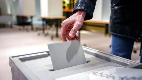 Eine Hand steckt einen Stimmzettel in eine Wahlurne. Die Person trägt eine schwarze Jacke. Im Hintergrund sind Tische und Wahlkabinen zu sehen. Ein Teil des Texts auf der Wahlurne ist sichtbar: „Abde...“.