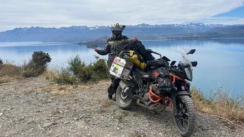 Tristan am Wasser in Chile Chico, Chile. Mit voller Motorradmontur und seiner bepackten Maschine steht der 22-Jährige vor einer Wasserfläche, im Hintergrund erhebt sich ein Bergpanorama mit schneebedeckten Gipfeln. 