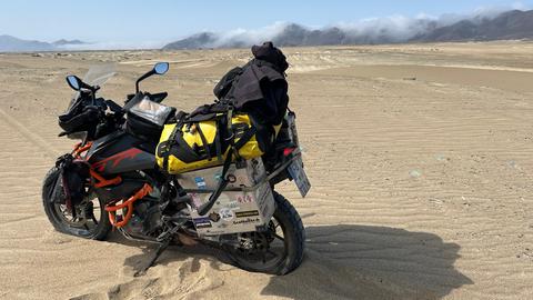 Tristan mit seinem Motorrad in Peru - die bepackte Maschine steht auf Wüstensand, der das Bild dominiert. Nur im Hintergrund sind Hügel zu erahnen. 