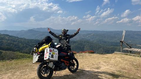 Tristan mit seinem Motorrad in Yang Moen, Provinz Chiang Mai, Thailand. In voller Motorradmontur steht der 22-Jährige mit ausgebreiteten Armen auf einer Anhöhe. Vor ihm sein Motorrad. Im Hintergrund die weite Landschaft mit bewaldeten Bergen. Am blauen Himmel helle Wolken.