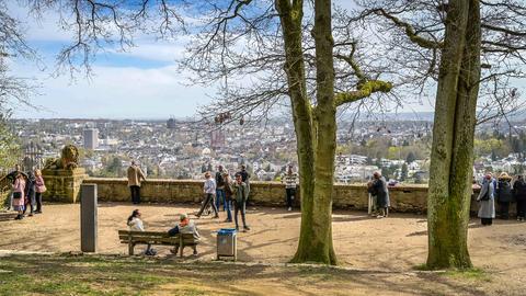 Blick von oben auf Wiesbaden von einer nahe gelegenen öffentlichen Terrasse, die am unteren, vorderen Bildrand zu sehen ist und auf welcher sich Besuchende tummeln.