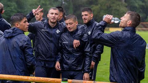 Einige Spieler des SV Darmstadt stehen im Regen auf dem Trainingsplatz. Filip Stojilković hat den Arm um Marvin Mehlem gelegt und macht mit seiner rechten Hand das Peace-Zeichen.