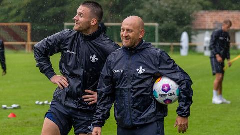 Darmstadt-Torhüter Marcel Schuhen stützt einen Arm in die Seite und lehnt sich an Trainer Torsten Lieberknecht, der einen Ball unter den Arm geklemmt hat und lacht.