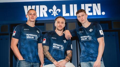 Drei Männer tragen das Sondertrikot des SV Darmstadt 98