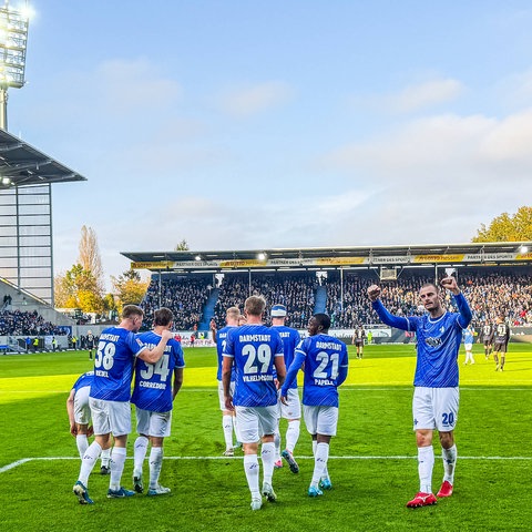 Eine Grupppe von Lilien-Spielern in weiß-blauen Trikots stehen auf einem Fußballfeld und jubeln.