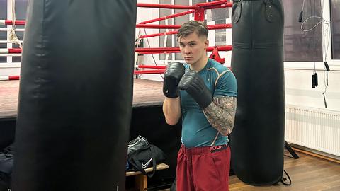 Ein Mann mit schwarzen Boxhandschuhen steht zwischen zwei dunklen Boxsäcken und vor einem Boxring und schaut in die Kamer.