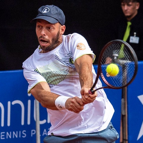 Ein Mann mit Tennisschläger in der Hand, in dessen Netz ein gelber Ball fliegt.