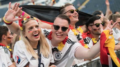 Zwei Menschen schauen in die Kamera, Deutschlandfahnen in der Hand, schwarz-rot-gold geschminkt. Sie stehen in einer Menschentraube, die beim Public Viewing einem Fußballspiel zuschaut.