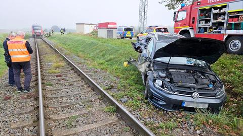 Ein beschädigtes Auto steht neben Bahngleisen, dahinter ein Feuerwehrwagen und daneben zwei Feuerwehrleute. Im Hintergrund in der Ferne ein Zug auf den Gleisen.