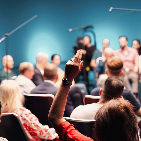 Viele Menschen sitzen zusammen. Im Vordergrund eine Hand, die sich meldet. Im Hintergrund unscharf eine Kamera und ein Mikrofon.