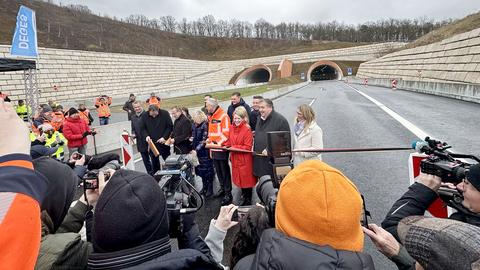 Mehrere Menschen schneiden ein schwarz-rot-goldenes Band durch, um das Teilstück der A44 zu eröffnen. Im Hintergrund sind zwei Tunnelröhren zu sehen.