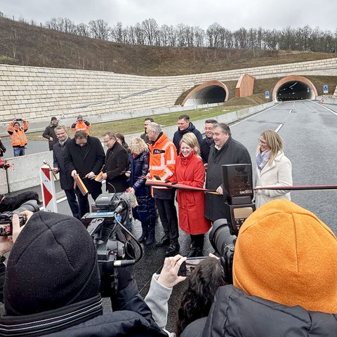 Mehrere Menschen schneiden ein schwarz-rot-goldenes Band durch, um das Teilstück der A44 zu eröffnen. Im Hintergrund sind zwei Tunnelröhren zu sehen.