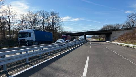 Eine zweispurige Autobahn unter einer Brücke. Auf der linken Spur fahren mehrere Fahrzeuge, darunter ein LKW mit dem Schriftzug. Die rechte Spur ist frei. Im Hintergrund sind Bäume und ein blauer Himmel mit einigen Wolken zu sehen. Auf der Brückenmauer befindet sich ein Graffiti.