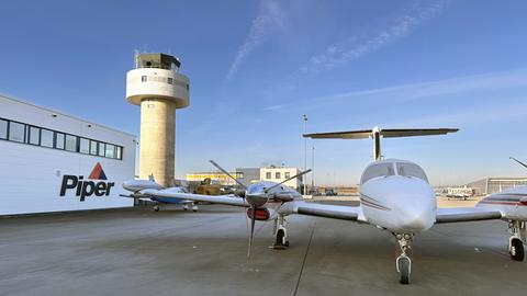 Ein kleines Flugzeug steht vor einem Gebäude. Am Giebel des Gebäudes ist die Aufschrift "Piper" zu lesen.