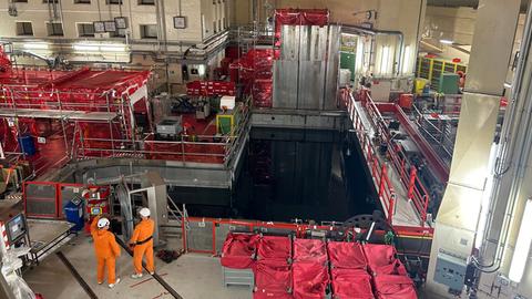 Zwei Männer in orangefarbenen Overalls stehen vor dem Abkühlbecken im Inneren des ehemaligen Atomkraftwerks Biblis, um sie herum hohe Betonwände und rot verpackte Pakete