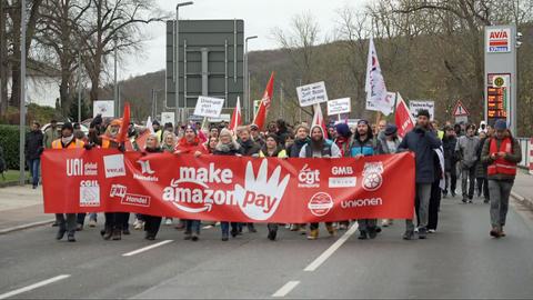 "Make Amazon Pay" ("Lasst Amazon bezahlen") steht auf einem Transparent, das so breit ist wie die Straße und von streikenden Amazon-Mitarbeitern getragen wird. Zu sehen sind auf dem Bild etwa 30 Menschen.