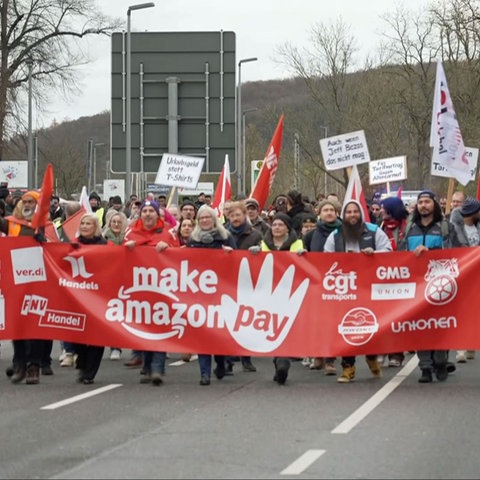 "Make Amazon Pay" ("Lasst Amazon bezahlen") steht auf einem Transparent, das so breit ist wie die Straße und von streikenden Amazon-Mitarbeitern getragen wird. Zu sehen sind auf dem Bild etwa 30 Menschen.