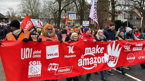 "Make Amazon Pay" ("Lasst Amazon bezahlen") steht auf einem Transparent, das so breit ist wie die Straße und von streikenden Amazon-Mitarbeitern getragen wird. Zu sehen sind auf dem Bild etwa 30 Menschen.