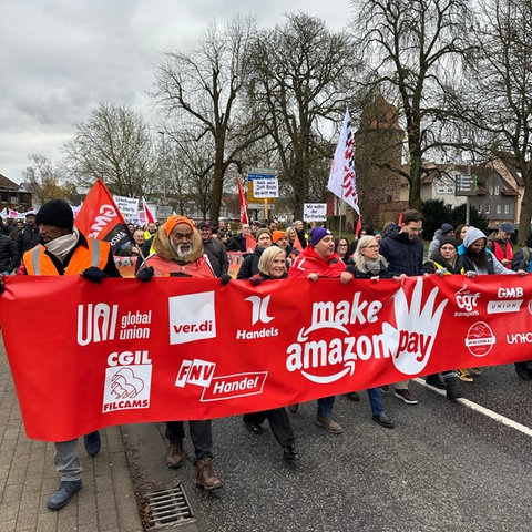 "Make Amazon Pay" ("Lasst Amazon bezahlen") steht auf einem Transparent, das so breit ist wie die Straße und von streikenden Amazon-Mitarbeitern getragen wird. Zu sehen sind auf dem Bild etwa 30 Menschen.