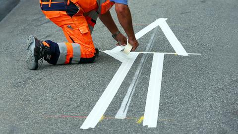 Klebestreifen, die einen Pfeil bilden, auf Asphalt. Daneben ein Arbeiter in neonfarbener Arbeitskleidung