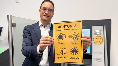 Ein Mann steht vor einem Geldautomaten, schaut in die Kamera und hält ein gelbes Schild in den Händen, auf welchem "Achtung! Verbaute Schutzmechanismen:" steht, darunter Piktogramme.