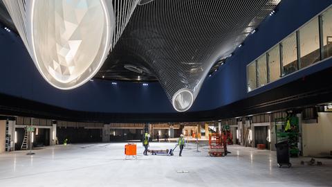 Bauarbeiter ziehen einen Palettenwagen durch die Markthalle des Terminal 3. Zu sehen sind futuristisch anmutende Deckengebilde. 