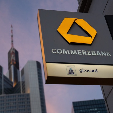 Im Bildvordergrund ein Commerzbank-Schild an einer Filiale in Frankfurt, im Hintergrund die Hochhäuser der Commerzbank.