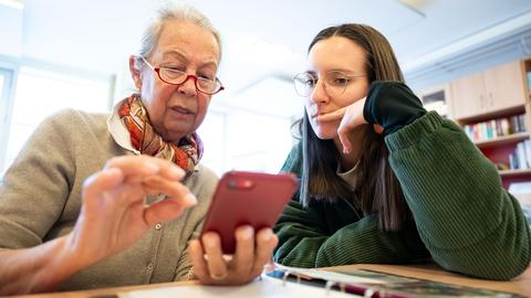 Eine ältere Frau und eine jüngere Frau sitzen an einem Tisch. Sie schauen gemeinsam auf das Display eines Smartphones.