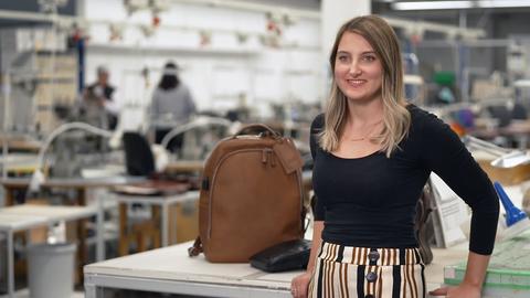 Eine Auszubildene zum Beruf der Feintäschnerin steht in der Fabrik, in der sie arbeitet. Hinter ihr sind Taschen un Rucksäcke zu sehen, die sie hergestellt hat.