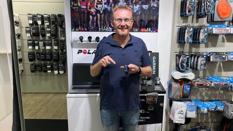 Ein Mann mit Brille und blauem Polo-Shirt steht in einem Laden vor einer Wand mit Laufschuhen und zeigt auf eine Smartwatch in seiner Hand