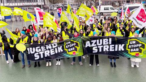 Streikende Azubis mit gelben Wanrwesten und roten Trillerpfeifen. Sie tragen Verdi-Fahnen und Plakate mit Aufschriften wie "Deine Mudda wird unterversorgt!".