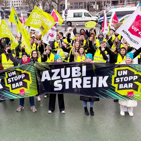 Streikende Azubis mit gelben Wanrwesten und roten Trillerpfeifen. Sie tragen Verdi-Fahnen und Plakate mit Aufschriften wie "Deine Mudda wird unterversorgt!".