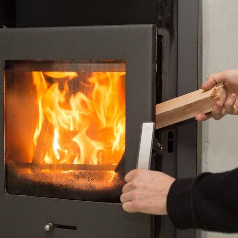 Ein Mann legt in seiner Wohnung ein Stück Holz in einen Kaminofen, in dem ein Feuer brennt. 