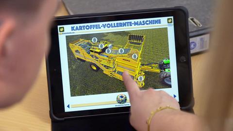 Nahaufnahme von einem Tablet, auf welchem eine landwirtschaftliche Großmaschine von oben mit Erklärtexten zu sehen ist. Oberdrüber steht "Kartoffel-Vollernte-Maschine". Ein Finger zeigt auf die Darstellung. Angeschnitten und unscharf zwei Köpfe am Rand des Bildes.